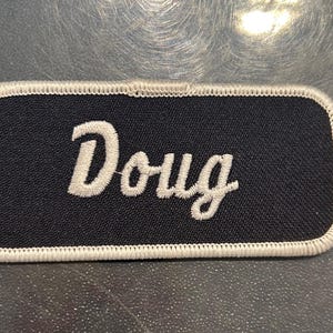 May include: A black fabric name tag with white stitching around the edges. The name "Doug" is embroidered in white on the tag.