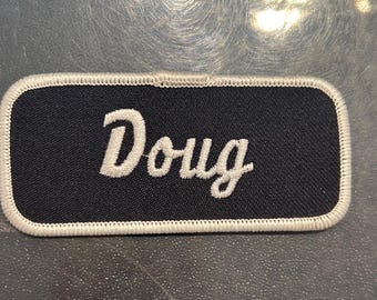 May include: A black fabric name tag with white stitching around the edges. The name "Doug" is embroidered in white on the tag.