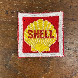 Op de afbeelding: Een rood en wit geborduurde patch met een gele schelp en het woord "SHELL" in rode letters.