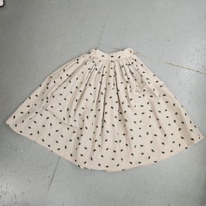 Puede incluir: Una falda de vichy blanca y marrón con un estampado de hojas verdes. La falda tiene una cintura fruncida y una silueta amplia y fluida.