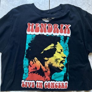 May include: Black t-shirt featuring a colorful graphic of Jimi Hendrix with the text "HENDRIX LIVE IN CONCERT". The shirt has a distressed look with cut details on the sleeves and neckline. The graphic has red, yellow, blue, and white colors.