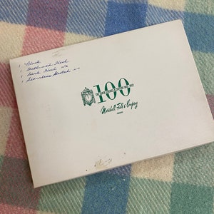 May include: A white box with green text that reads "The Clock Strikes a Century 1852-1952" and "Marshall Field & Company Chicago". The number "100" is printed in green on the box. The box has handwritten notes on the top that read "1 Black Outlined Heel Dark Neck 1012 1 Seamless Stretch 10-11"