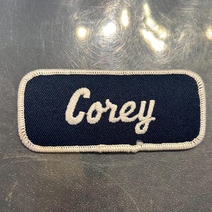 May include: A blue fabric patch with white stitching around the edges. The patch has the name "Corey" embroidered in white on a dark blue background.