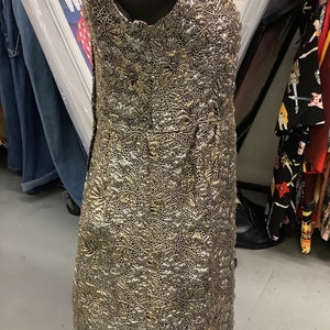 May include: A sleeveless gold and silver dress with a textured, floral pattern. The dress is on a mannequin and is displayed on a metal stand.