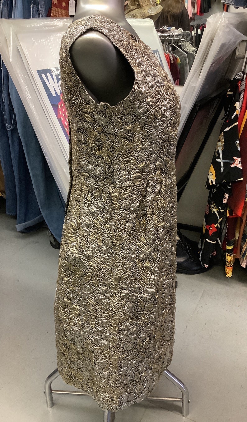 May include: A sleeveless gold and silver dress with a textured, floral pattern. The dress is on a mannequin and is displayed on a metal stand.