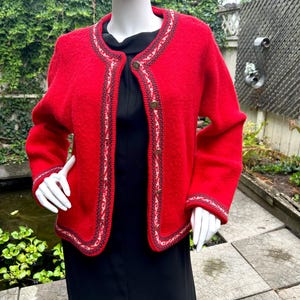 May include: A bright red cardigan with a decorative trim in white, black, and red. The cardigan has long sleeves and is fastened with gold-colored buttons. The garment is displayed on a mannequin, showcasing its design and fit.