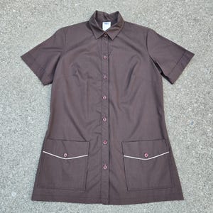 Puede incluir: Camisa marrón de manga corta con botones y dos bolsillos delanteros. Los bolsillos y el cuello tienen ribetes blancos. La camisa tiene un cuello clásico y está hecha de una tela lisa.