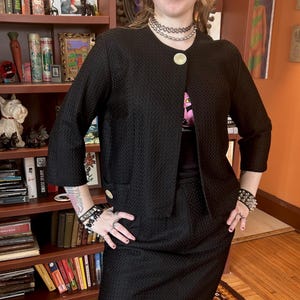 Peut inclure: Ensemble jupe-veste noire texturée. La veste a des manches trois-quarts et de gros boutons dorés. L'ensemble est porté sur un t-shirt noir. L'ensemble est de style vintage.