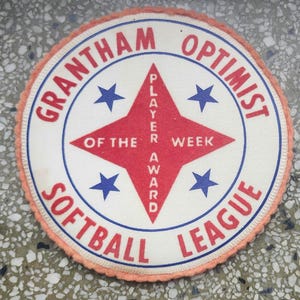 Può includere: Una toppa circolare con la scritta "GRANTHAM OPTIMIST SOFTBALL LEAGUE" in rosso e blu. Il centro presenta un'esplosione di stelle rossa con "PLAYER OF THE WEEK AWARD" in bianco. Quattro stelle blu circondano l'esplosione.