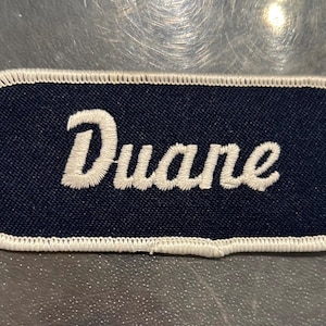 May include: A white embroidered name tag with a navy blue background. The name "Duane" is stitched in white on the tag.
