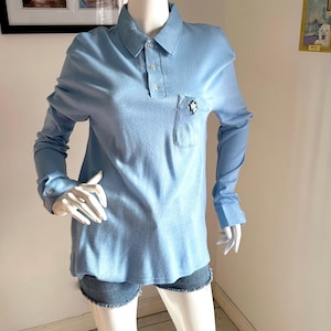 Puede incluir: Polo de manga larga azul claro con un bolsillo en el pecho. La camisa tiene un cuello clásico y puños abotonados. Un pequeño emblema en blanco y negro está en el bolsillo. La camisa se combina con pantalones cortos de mezclilla.