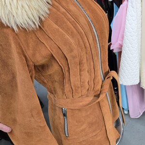 May include: A brown suede jacket with a cream-colored faux fur collar. The jacket features a vertical zipper, a belt, and a small zippered pocket. The jacket has a textured, ribbed design on the front.
