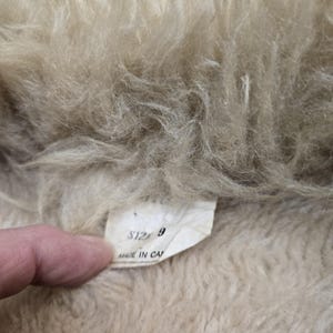 May include: Close-up of a beige, fluffy textile with a tag. The tag reads "SIZE 9" and "MADE IN CA". The textile has a soft, textured appearance, with visible fibers and a light color.