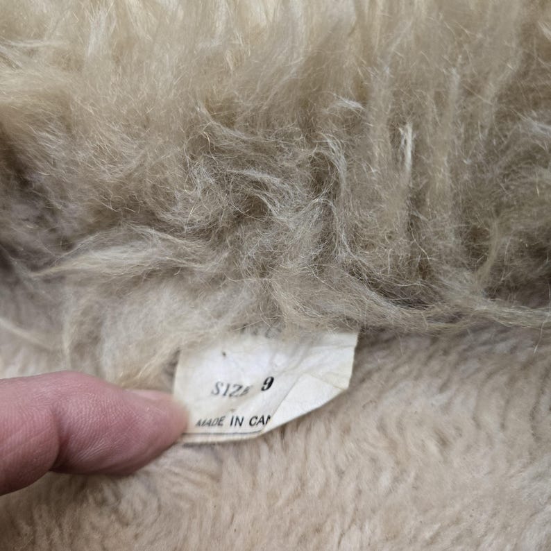 May include: Close-up of a beige, fluffy textile with a tag. The tag reads "SIZE 9" and "MADE IN CA". The textile has a soft, textured appearance, with visible fibers and a light color.