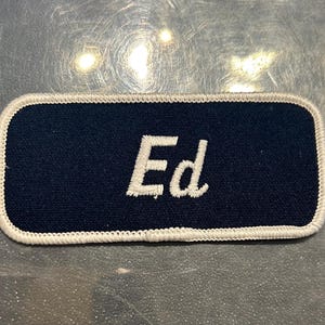 May include: A black fabric patch with white stitching around the edges. The patch has the name "Ed" embroidered in white thread.