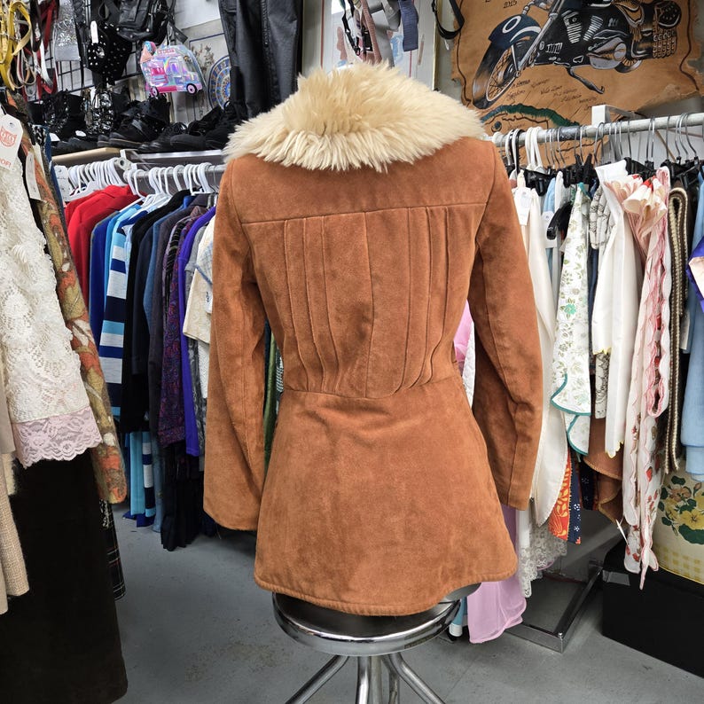 May include: A brown suede jacket with a cream-colored faux fur collar. The jacket has long sleeves and a fitted waist, with vertical pleats on the back. The jacket is displayed on a metal stool in a vintage clothing store.