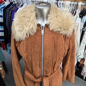 May include: A brown suede jacket with a faux fur collar and a belt. The jacket has a zipper closure and vertical stitched details. The jacket is displayed on a mannequin. Clothing.