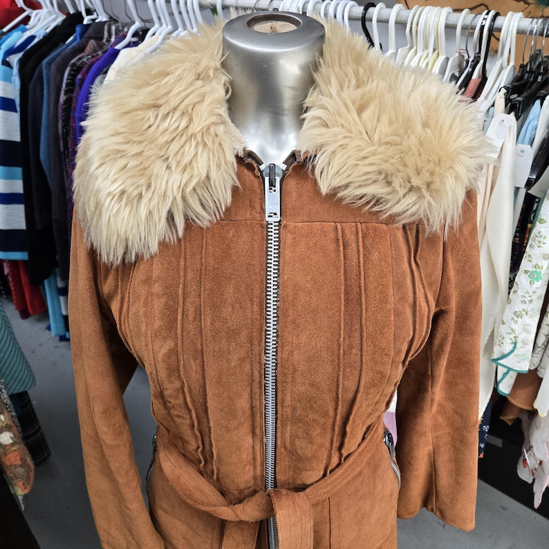 May include: A brown suede jacket with a faux fur collar and a belt. The jacket has a zipper closure and vertical stitched details. The jacket is displayed on a mannequin. Clothing.