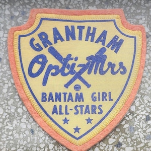 May include: A yellow and orange felt patch with the text "Grantham Optimists Bantam Girl All-Stars" and three blue stars.