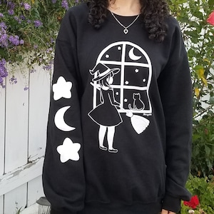 Puede incluir: Sudadera negra con un gráfico blanco de una bruja mirando por una ventana con un gato. Las mangas tienen estrellas blancas y una luna creciente.