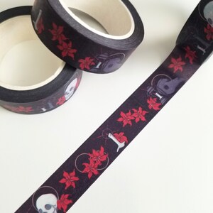 May include: Black and red washi tape with a repeating pattern of skulls, flowers, and a skeletal hand holding a candle. The tape is rolled up and partially unrolled, showing the pattern.