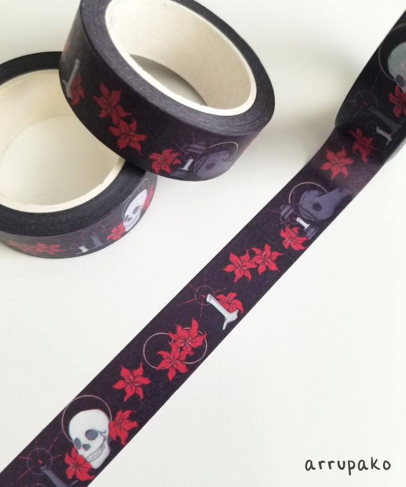 May include: Black and red washi tape with a repeating pattern of skulls, flowers, and a skeletal hand holding a candle. The tape is rolled up and partially unrolled, showing the pattern.