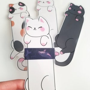 May include: Three cat-shaped bookmarks, one white, one black, and one calico. The white bookmark has a whale sticker on it.