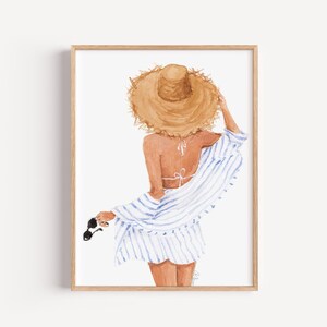 May include: Watercolour print of a person wearing a straw hat, a white and blue striped cover-up, and holding sunglasses. The artwork is framed in a light brown wooden frame. The person is facing away from the viewer.