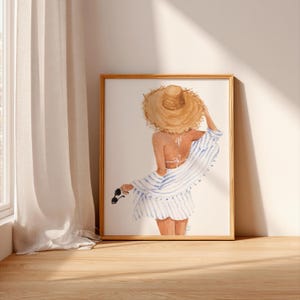 May include: Framed watercolour print. The artwork shows a person wearing a straw hat, a white and blue striped cover-up, and a white swimsuit. The person is holding sunglasses. The art is displayed on a wall with a wooden floor.