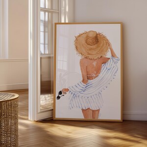 May include: Watercolour print of a person wearing a straw hat, a white bikini top, and a blue and white striped cover-up. The artwork is framed in a light wood frame and leans against a white wall. Sunglasses are held in the hand.