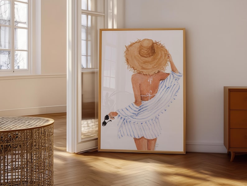 May include: Watercolour print of a person wearing a straw hat, a white bikini top, and a blue and white striped cover-up. The artwork is framed in a light wood frame and leans against a white wall. Sunglasses are held in the hand.