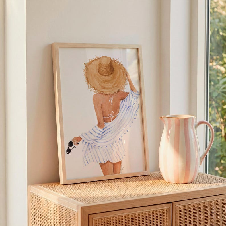 May include: A framed watercolour print shows a person in a straw hat and striped cover-up, holding sunglasses. A pink and white striped jug sits on a wooden cabinet. The artwork has a beach theme.