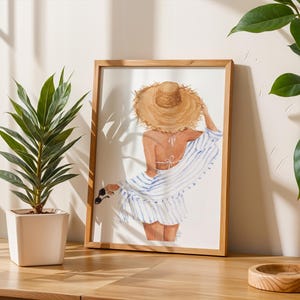 May include: A framed watercolour print showing a woman in a straw hat and striped cover-up, viewed from behind. Sunglasses are held in her hand. A potted plant and wooden tray are on a wooden surface.