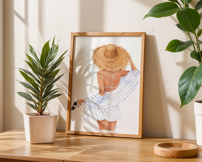 May include: A framed watercolour print showing a woman in a straw hat and striped cover-up, viewed from behind. Sunglasses are held in her hand. A potted plant and wooden tray are on a wooden surface.