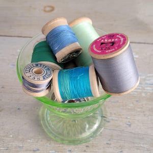 May include: A collection of spools of thread in various colors, including green, blue, teal, and gray, are displayed in a clear green glass vase. The spools are made of wood and have labels with text.