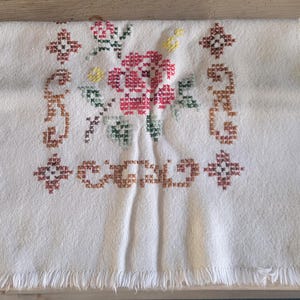 May include: White cotton table runner with a red, green, and yellow embroidered floral design. The design features a large flower in the center with smaller flowers and leaves surrounding it.