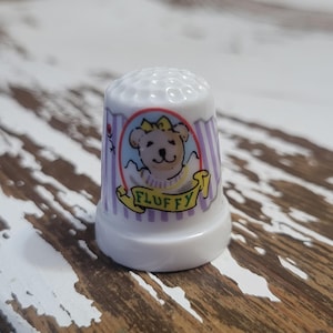 May include: White ceramic thimble with a purple and white striped design. A cartoon bear with a yellow bow is in the center of the thimble with the word "FLUFFY" written on a yellow banner.