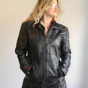Puede incluir: Una chaqueta de cuero negro con diseño de patchwork y costuras visibles, cremallera frontal completa, cuello puntiagudo y mangas largas. El forro interior es rojo, visible en el cuello y los puños, y la chaqueta tiene dos bolsillos delanteros.