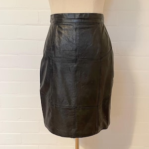 May include: A black leather skirt with a patchwork design. The skirt is knee-length and has a high waist. The leather appears to be slightly textured. The skirt is displayed on a mannequin against a white brick wall.