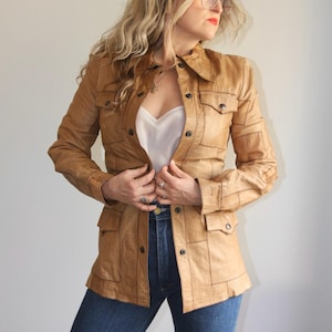May include: A tan leather jacket constructed from multiple rectangular panels with a pointed collar, long sleeves, and four front pockets with button closures. The jacket features a central button-down placket and visible stitching along the seams.