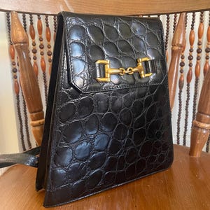 Può includere: Borsa a mano in pelle nera con motivo a rilievo effetto coccodrillo. La borsa ha una forma rettangolare con chiusura a patta assicurata da una chiusura e catena in metallo dorato. La borsa è su una sedia di legno.