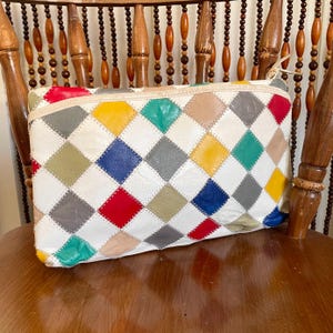 May include: A multi-coloured patchwork leather clutch bag with a zip. The bag has a diamond pattern in various colours, including red, yellow, blue, green, and grey, set against a white background. The clutch is on a wooden chair.