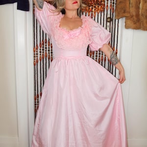 May include: A light pink vintage dress with a lace-trimmed neckline and puffed sleeves. The dress features a fitted bodice and a flowing, floor-length skirt. A pink bow accents the bodice. The dress is made of a shiny, silky material.