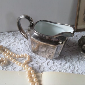 Könnte beinhalten: Eine silberne Kanne mit weißem Innenraum, die auf einer weißen Spitzentischdecke steht. Eine Perlenkette liegt ebenfalls auf der Tischdecke.
