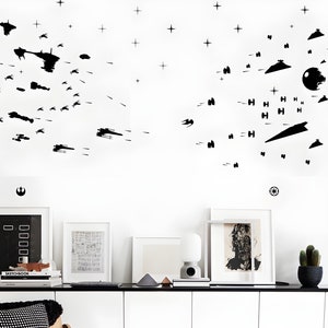 May include: A white wall with black Star Wars spaceship silhouettes and stars. The wall is decorated with a black Rebel Alliance symbol and a black Imperial symbol. There are three framed pictures and a lamp on a white shelf below the wall.