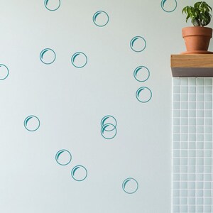 Puede incluir: Calcomanías de pared de burbujas azul turquesa en una pared azul claro. Las calcomanías son de varios tamaños y están dispuestas en un patrón aleatorio. Hay un estante de madera con una planta en maceta y dos botellas de jabón de manos encima.