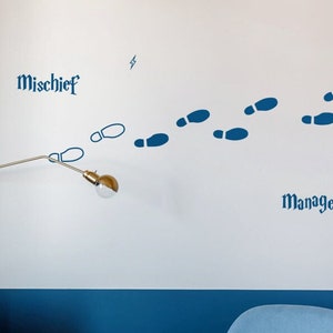 Puede incluir: Un vinilo decorativo de pared con huellas azules, un rayo y las palabras "Mischief Managed". También se ve una lámpara de pared dorada con una bombilla redonda. La habitación tiene una combinación de colores azul y blanco.