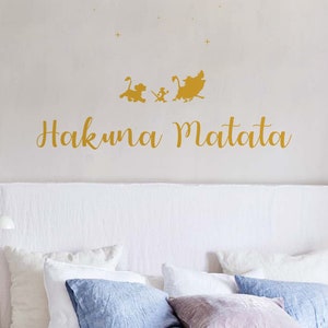 Peut inclure: Un autocollant mural doré représentant les personnages Simba, Timon et Pumbaa du film Disney Le Roi Lion. L'autocollant comprend également la phrase "Hakuna Matata" en écriture cursive.