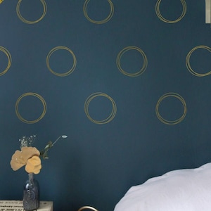 Può includere: Un muro blu-verde con adesivi circolari dorati. Il muro è decorato con un letto con lenzuola bianche e un piccolo vaso con fiori gialli su un tavolo di legno.