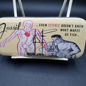 May include: A rectangular, cream-colored tin with the word "Fossil" in black script. The tin features a red and white anatomical illustration of a human figure and a scientist looking through a microscope. Text reads: "...EVEN SCIENCE DOESN'T KNOW WHAT MAKES US TICK."